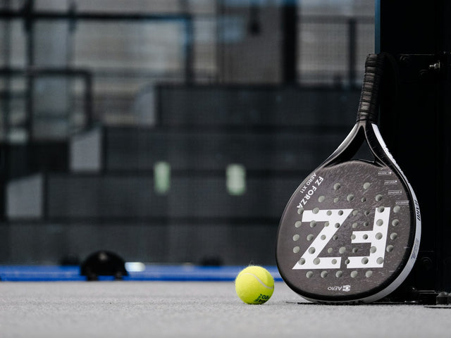 Raquette de padel posée debout à côté d'une balle jaune sur un court